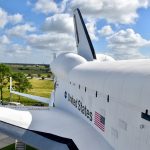 Looking down the length of a white Space Shuttle from a high platform. United States + flag printed on side of shuttle. View of fields and cloudy sky behind. You cannot see the nose of the shuttle in the photo