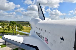 Looking down the length of a white Space Shuttle from a high platform. United States + flag printed on side of shuttle. View of fields and cloudy sky behind. You cannot see the nose of the shuttle in the photo