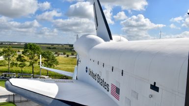Looking down the length of a white Space Shuttle from a high platform. United States + flag printed on side of shuttle. View of fields and cloudy sky behind. You cannot see the nose of the shuttle in the photo