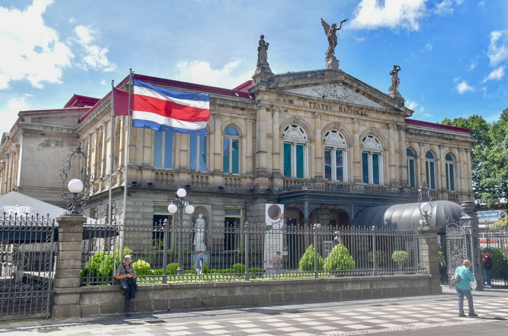 Front of the National Theatre in San Jose, Costa Rica. Large stone building with sculptures on the roof and black/white checked tiles in front. Costa Rica flag flying the the left
