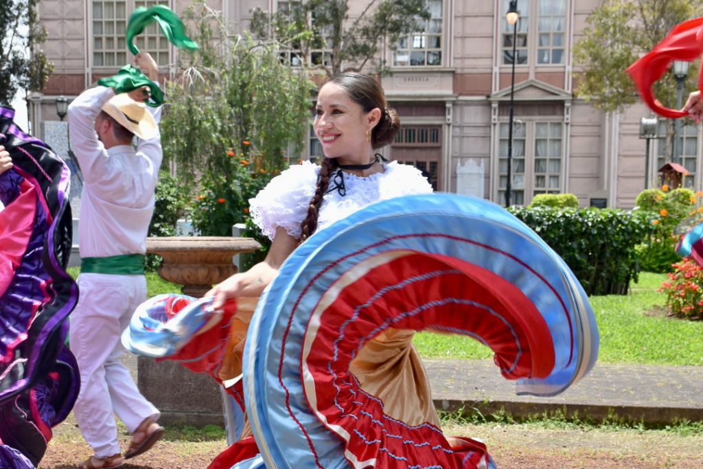 Beautiful smiling lady with brown hair back in a bun wearing Costa Rican traditional dress - white top, scarf around neck and blue / red skirt which is twirling whilst she dances in park. Make dancer in white behind her