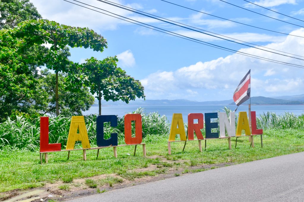 View of multicoloured sign by side of the road saying Laco Arenal (red, yellow, blue and white letters). Costa Rica flag at the end. Road in the foreground, lake behind with hills in background and a pale blue sky with white cloud