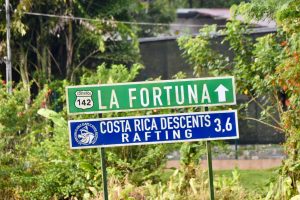 Green road sign to La Fortuna with white writing and arrow. Above a blue sign towards Raft activities. Background of tropical trees