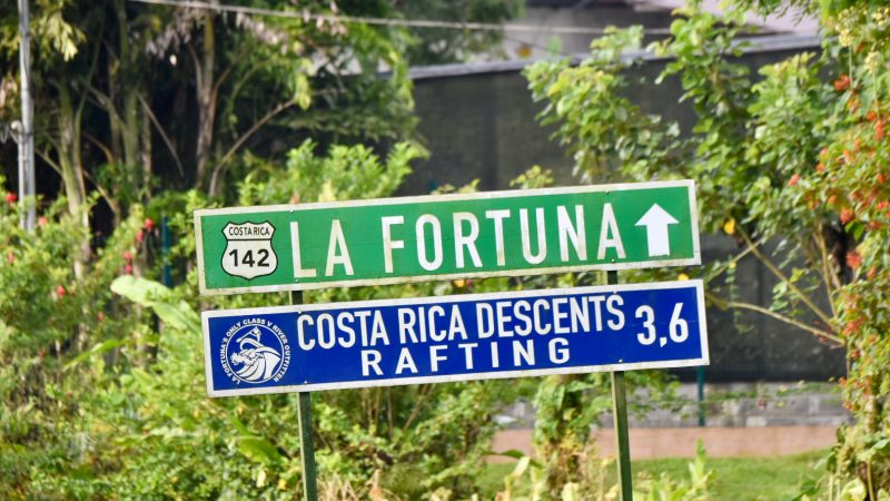 Green road sign to La Fortuna with white writing and arrow. Above a blue sign towards Raft activities. Background of tropical trees