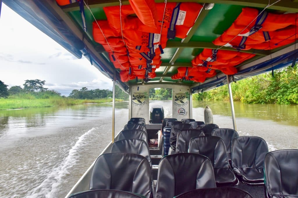 View down the length of an open sided boat travelling along a river with greenery on the river banks. There are red life jackets across the roof of the boat. No passengers in the photo.