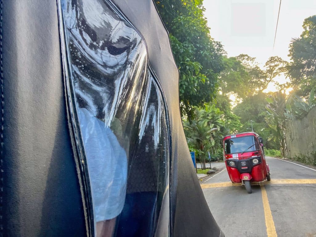 Photo taken from the back of a tuk tuk. View is looking back down to road to another tuk tuk taxi following behind. It is red