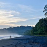 Sunrise photo of a black beach (volcanic sand) and sun coming through clouds illuminating a jogger on the beach and the waves