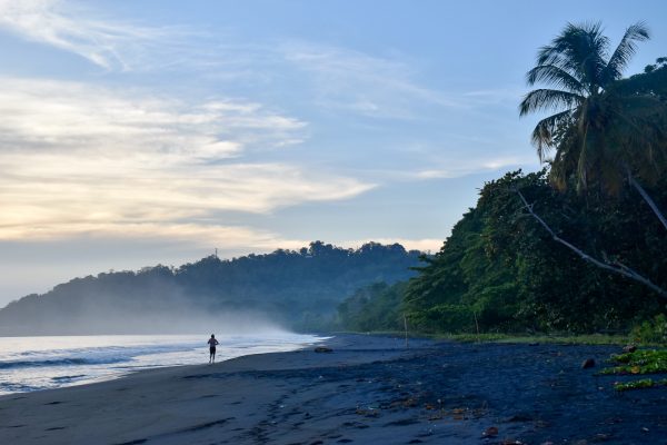 Sunrise photo of a black beach (volcanic sand) and sun coming through clouds illuminating a jogger on the beach and the waves