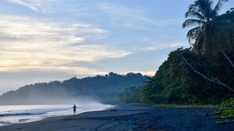 Sunrise photo of a black beach (volcanic sand) and sun coming through clouds illuminating a jogger on the beach and the waves