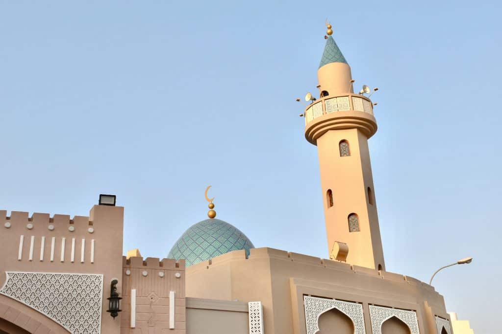 Creamy coloured Arabic buildings with a turquoise cupola and minaret, against pale blue sky