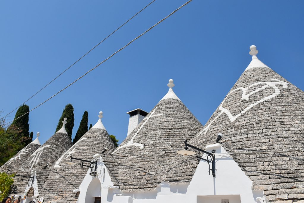 Row of 3 white washed stone buildings with grey tiled roofs each with a white symbol painted on. Blue sky in background and 2 power cables across top left of photo