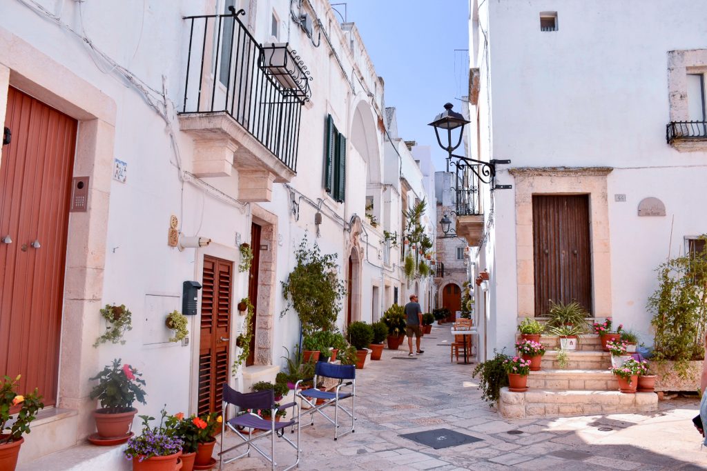 View of white washed houses on either side of a narrow alley in the sunshine. The houses have wooden doors, iron balconies and pots of plants on doorsteps