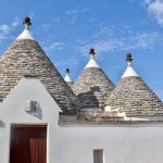 White washed stone building with three grey conical roofs against a blue sky. Dark brown door at the front of the property