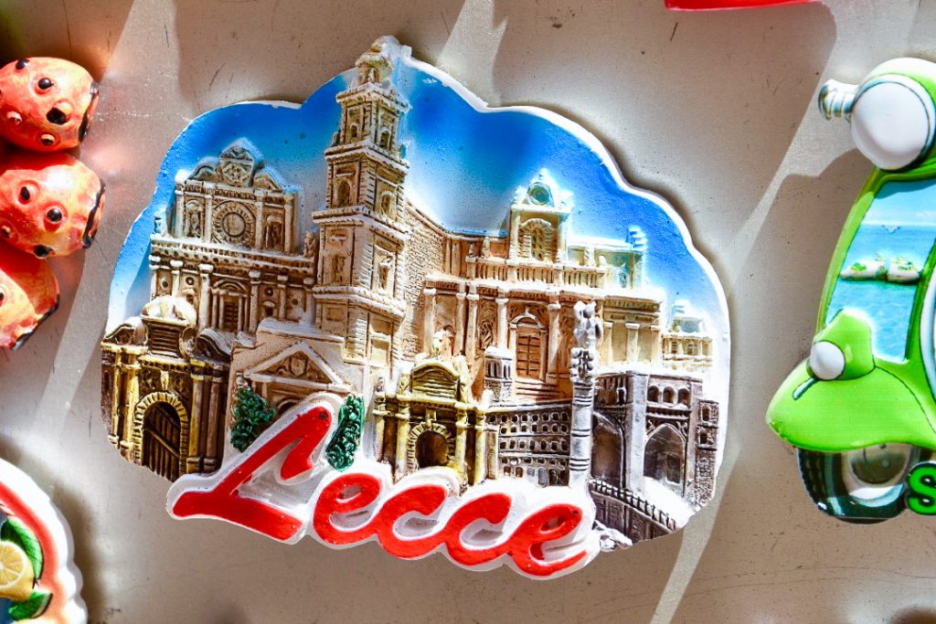 Oval fridge magnet featuring sandstone buildings of an Italian town against a blue sky and the name Lecce in red at the bottom
