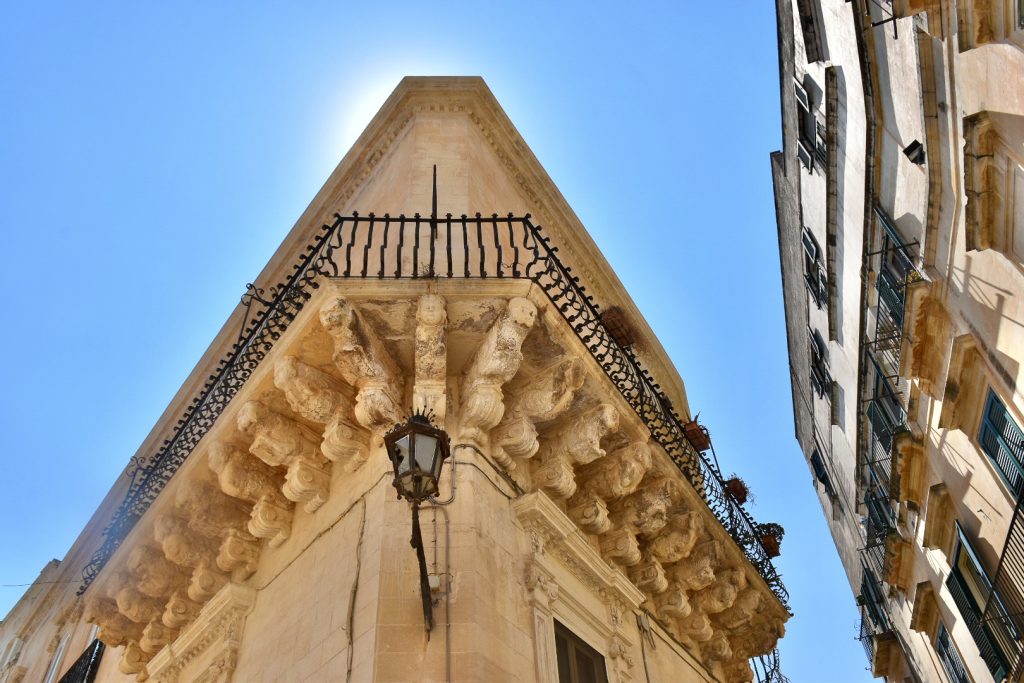 Photo of corner of sandstone building, taken from street and looking up towards the roof. Baroque carvings on the upper level of building. Blue sky behind