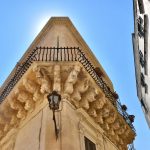 Photo of corner of sandstone building, taken from street and looking up towards the roof. Baroque carvings on the upper level of building. Blue sky behind
