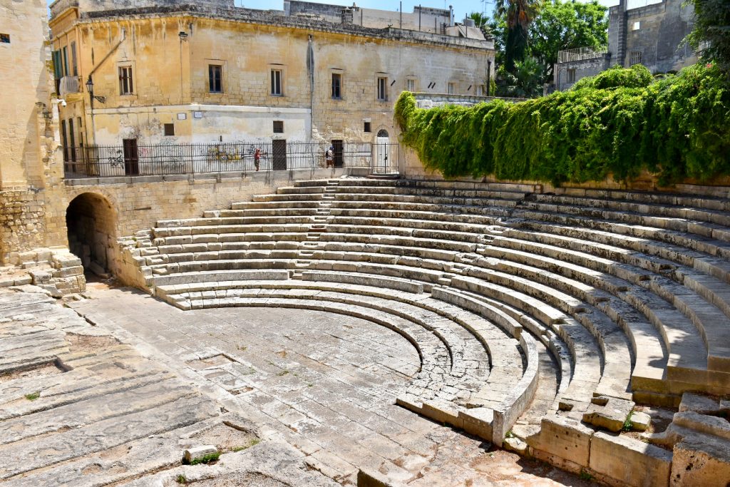 View of a semi-circular tier of seating in front of ocre coloured buildings and some shrubs. The seating forms the remains of a Roman Theatre