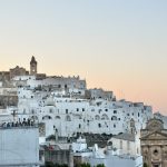 Old fashioned hilltop town with pink and orange sky behind it at dusk. Jumble of white and sand coloured houses on the hill
