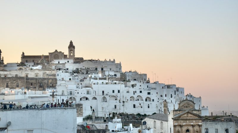 Old fashioned hilltop town with pink and orange sky behind it at dusk. Jumble of white and sand coloured houses on the hill