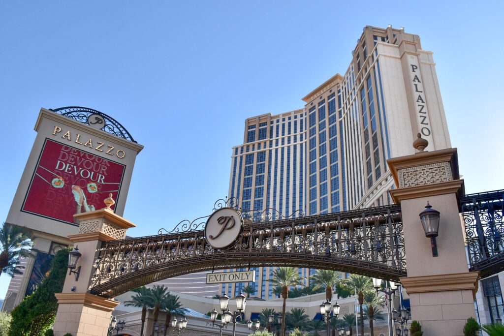 Looking up at a tall white modern hotel with blue windows. It has the word PALAZZO running down the right hand side. Across the bottom is an iron like bridge with a sign saying Exit Only on it. To the left a Sign for the Palazzo Hotel. Palm trees at the bottom. Pale blue sky above