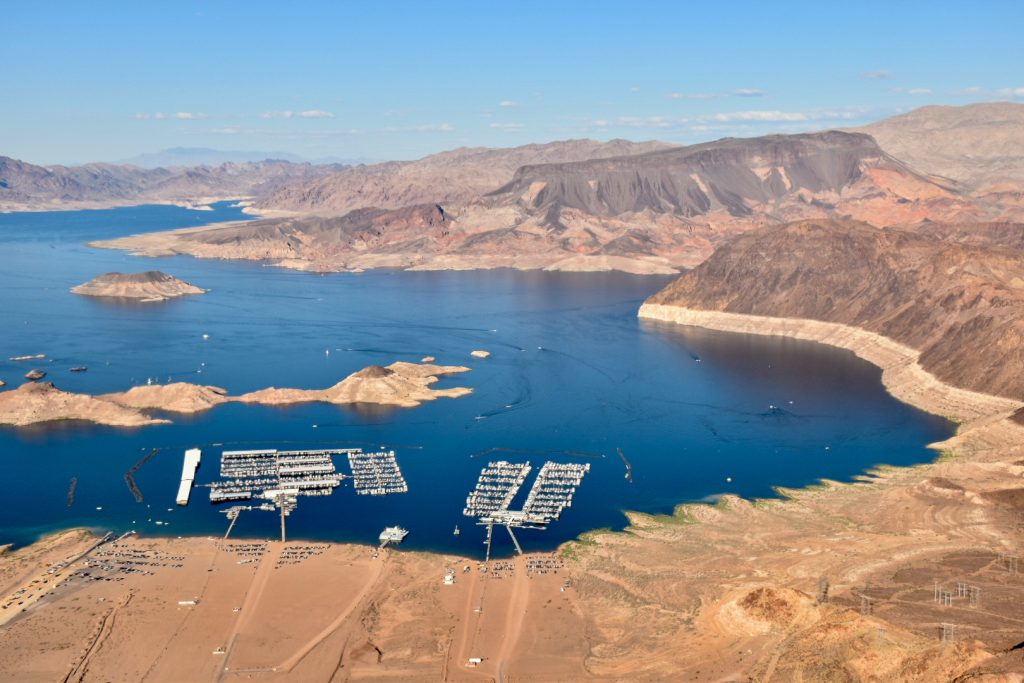 View from the air of a dark blue lake surrounded by golden rocky desert landscape. Some boat docks built into the water. Pale blue sky above