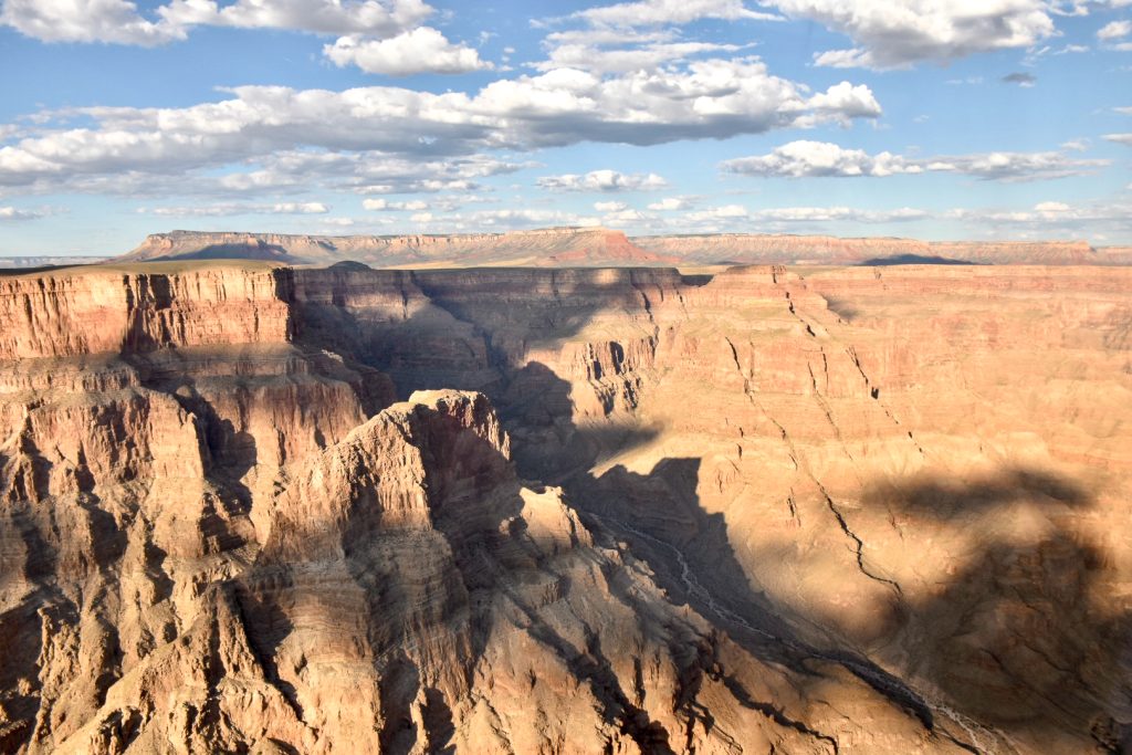 Pale blue sky and white fluffy clouds in top third of photo. Golden rocky canyon fills the other two-thirds of the photo. The rocks are striped and have shadows highlighting the different layers of the canyon