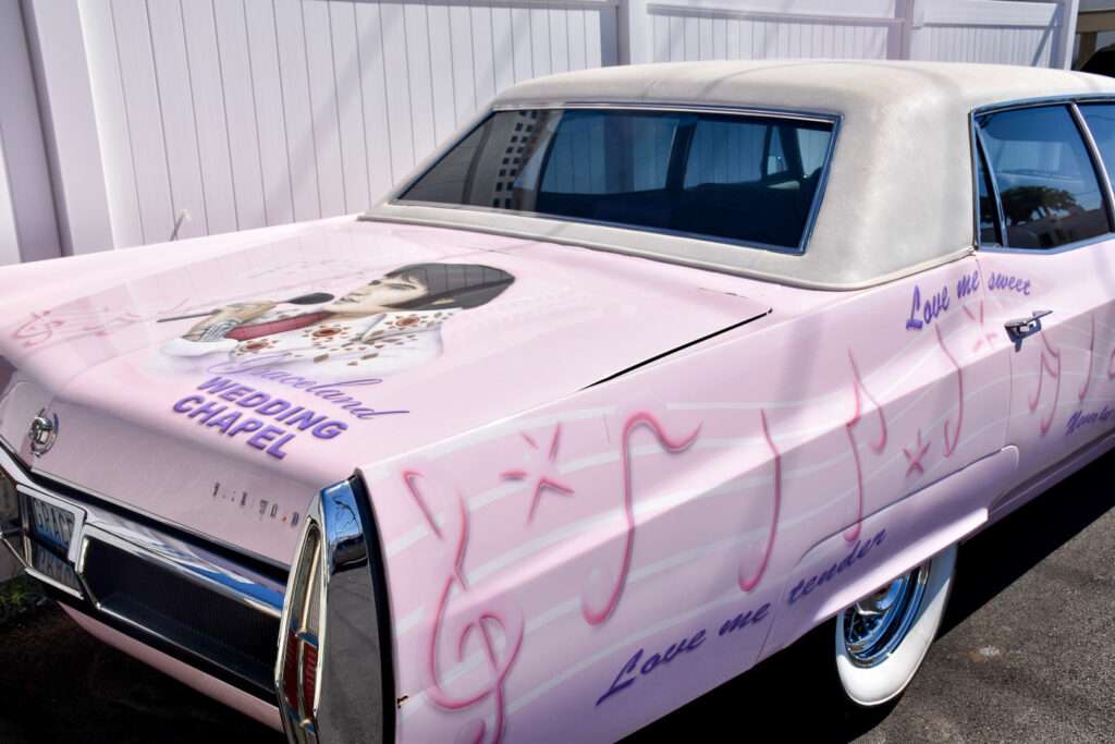 A pink limo is parked across the middle of the frame. It has a white roof and has a picture of Elvis on the trunk. It says Wedding Chapel.
