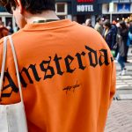 Back of man's shoulders wearing bright orange sweatshirt with word Amsterdam written in large black type face. White tote bag over shoulder