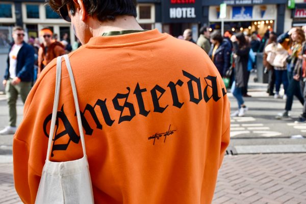 Back of man's shoulders wearing bright orange sweatshirt with word Amsterdam written in large black type face. White tote bag over shoulder