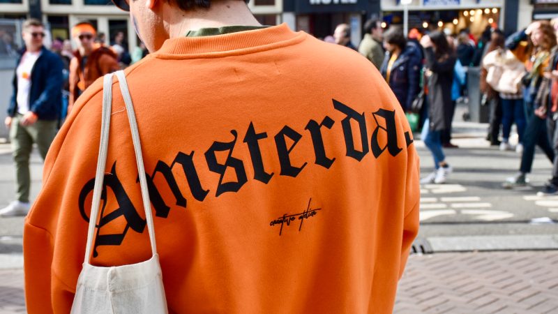 Back of man's shoulders wearing bright orange sweatshirt with word Amsterdam written in large black type face. White tote bag over shoulder