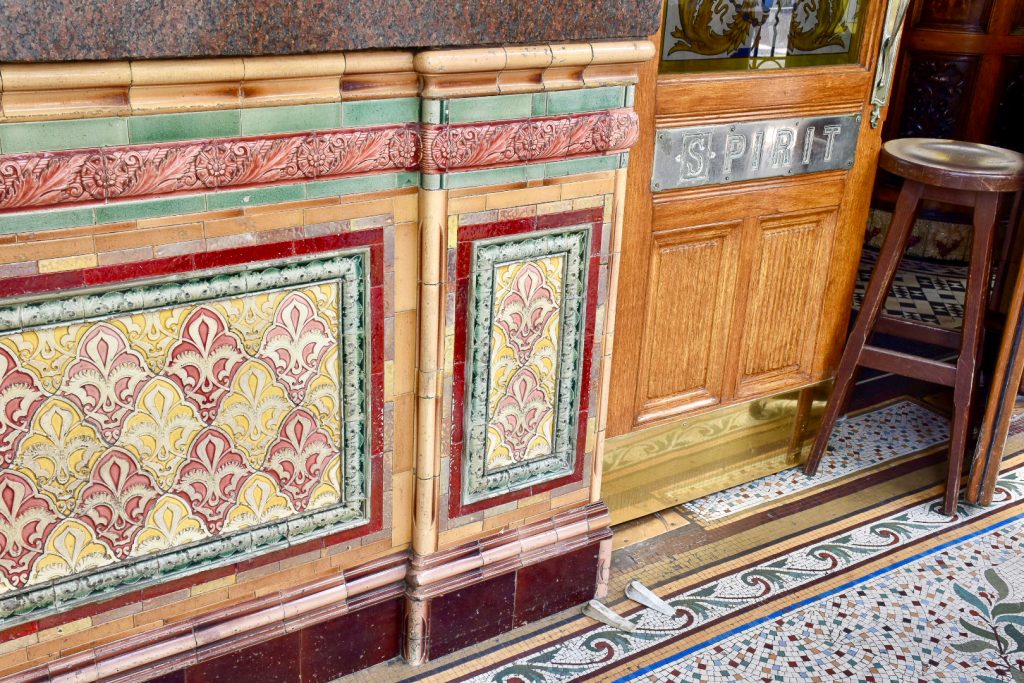 Close up of multicoloured tiled wall on the outside of a pub. Door to the right hand side. Plaque stating Saloon next to the door