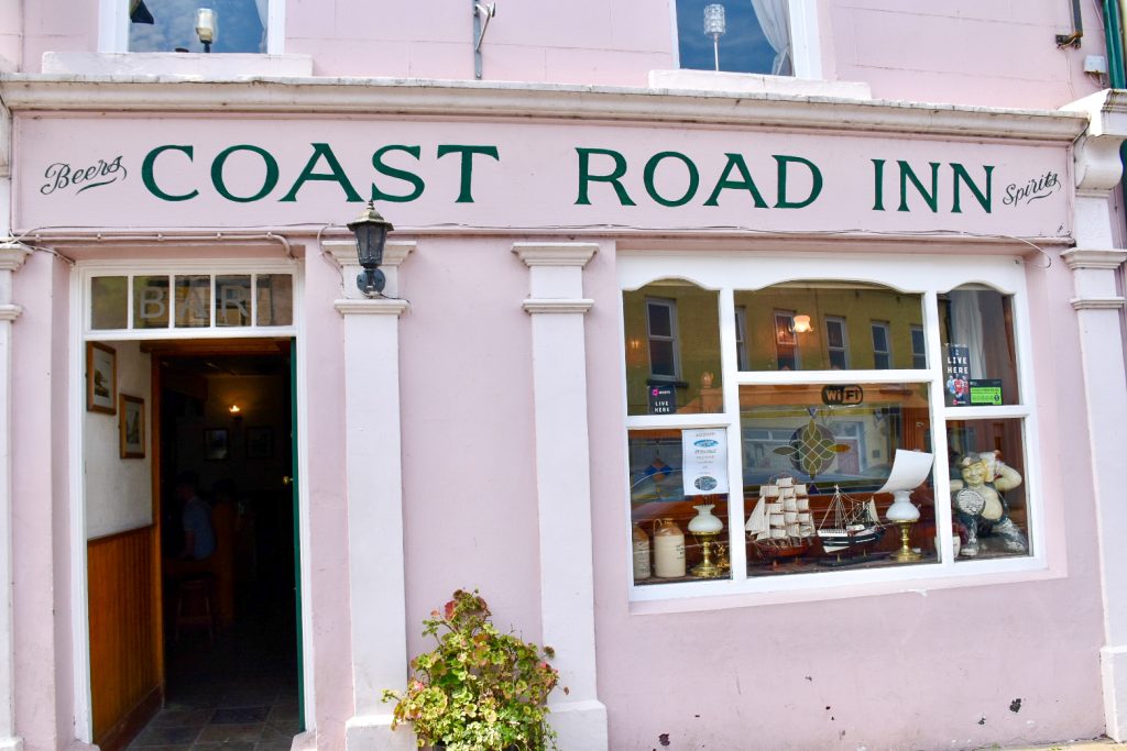 Front of a pink painted pub - open door on the left and window on the right. The Coast Road Inn painted above the door