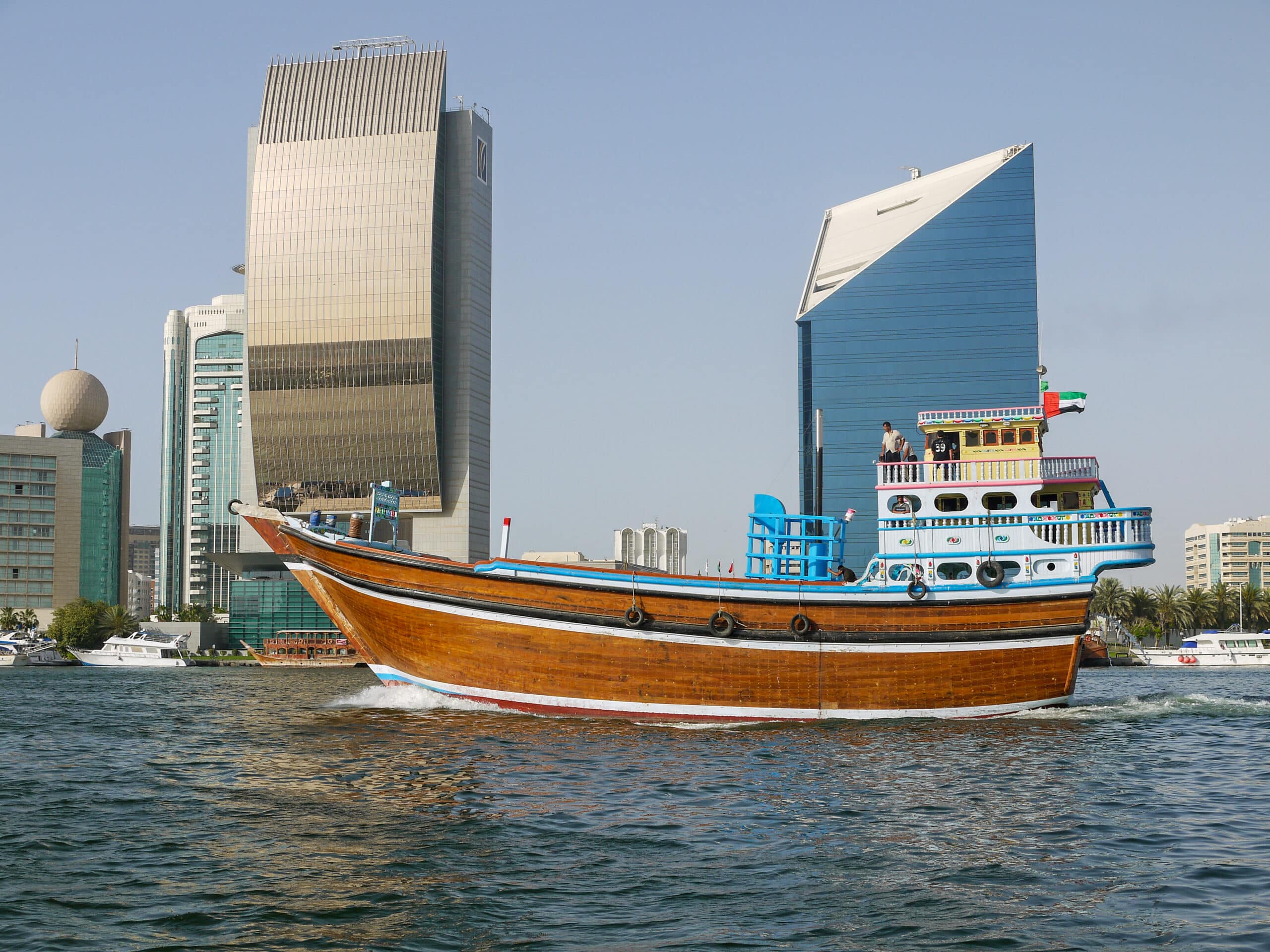 Traditional brown wooden boat sailing on river in front of modern skyscrapers - one gold and one blue