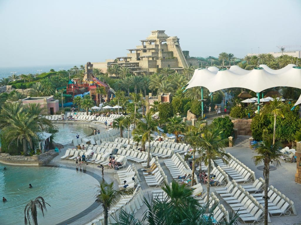 Waterpark with pool in foreground surrounded by sun loungers, palm trees and tall pyramid style water slide on horizon