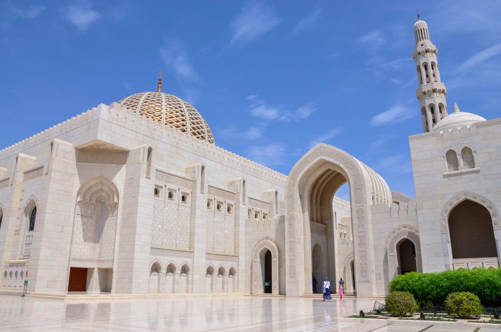 White marble buildings of a mosque. Gold cupola on the left hand square building. Large archway and minaret to the right hand side