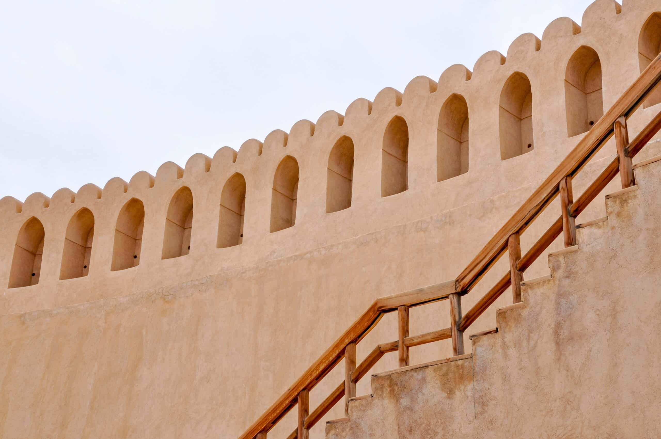 Photo of orange/brown wall of a fort. Looking upwards to the crenelated battlements. Wooden staircase from floor to top of battlements.