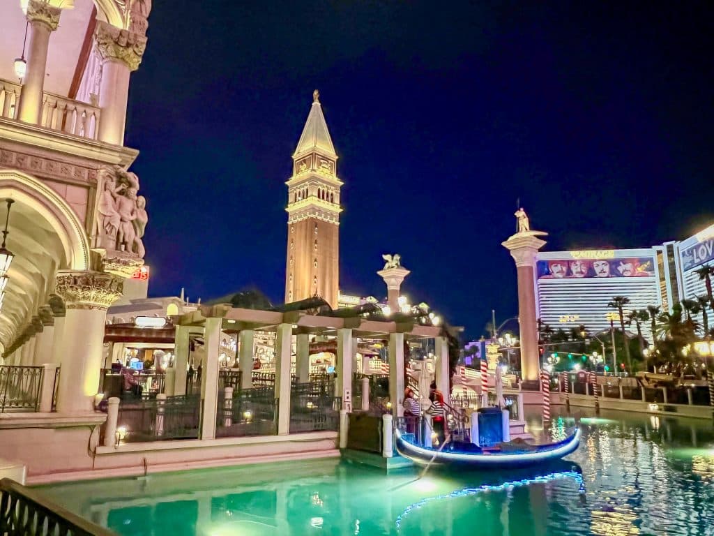 Night sky with illuminated buildings in front - tower and arced buildings behind a green canal with blue gondola boat on the water