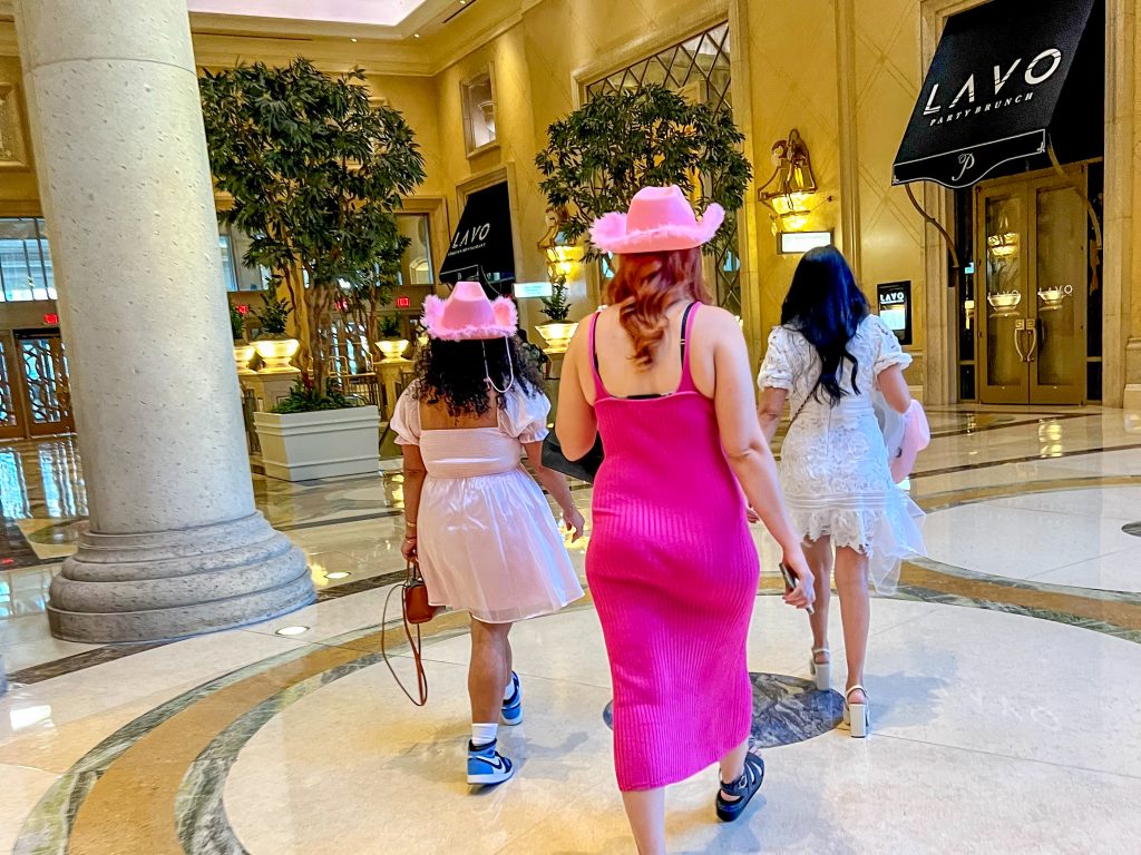 Three ladies walking away from the camera towards the door of a hotel lobby. The one closest to the camera is wearing a bright pink dress and pink cowboy hat, the lady on the left has a pale pink dress and cowboy had and the lady on the right has a white dress but no hat