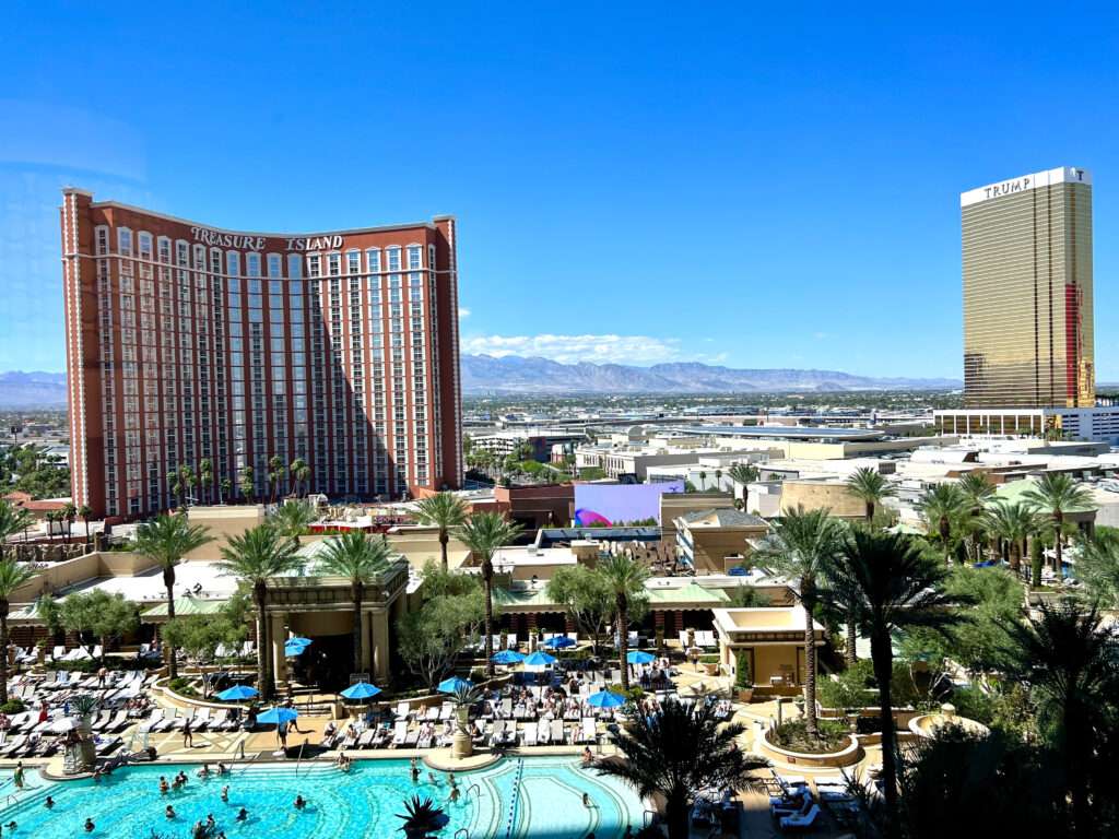 Hotel swimming pool, palm trees and loungers in the foreground. A large brown multi-storey hotel to the middle right hand side. Says Treasure Island on it. Desert town stretching beyond to the right. Blue sky above