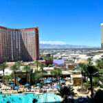 Hotel swimming pool, palm trees and loungers in the foreground. A large brown multi-storey hotel to the middle right hand side. Says Treasure Island on it. Desert town stretching beyond to the right. Blue sky above