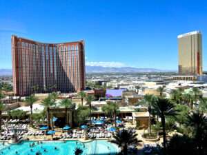 Hotel swimming pool, palm trees and loungers in the foreground. A large brown multi-storey hotel to the middle right hand side. Says Treasure Island on it. Desert town stretching beyond to the right. Blue sky above