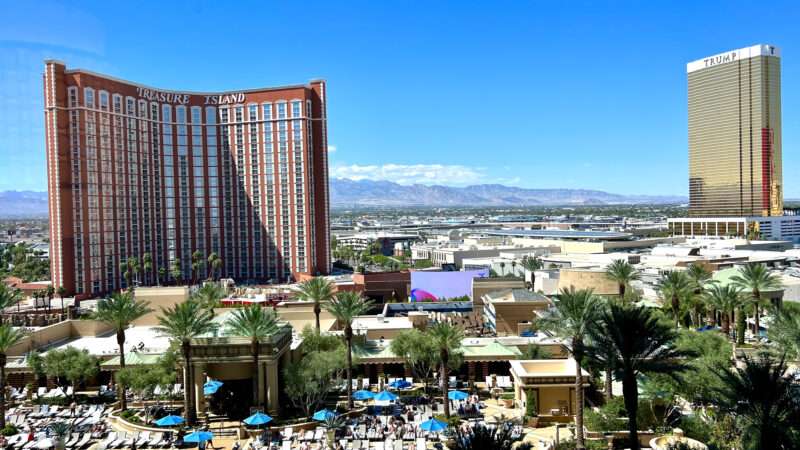 Hotel swimming pool, palm trees and loungers in the foreground. A large brown multi-storey hotel to the middle right hand side. Says Treasure Island on it. Desert town stretching beyond to the right. Blue sky above