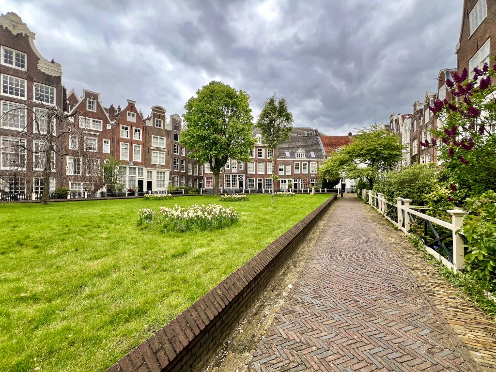 Looking down. path to the right hand side, grass verge to the left. Dutch brown brick buildings along the sides and back of the courtyard. Cloudy sky above.