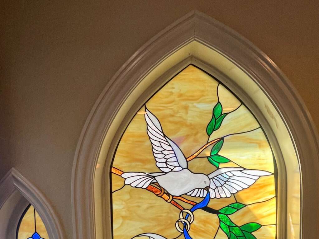 A stained glass window sits to the right hand side of the frame. It features a white dove sitting on a branch with leaves.