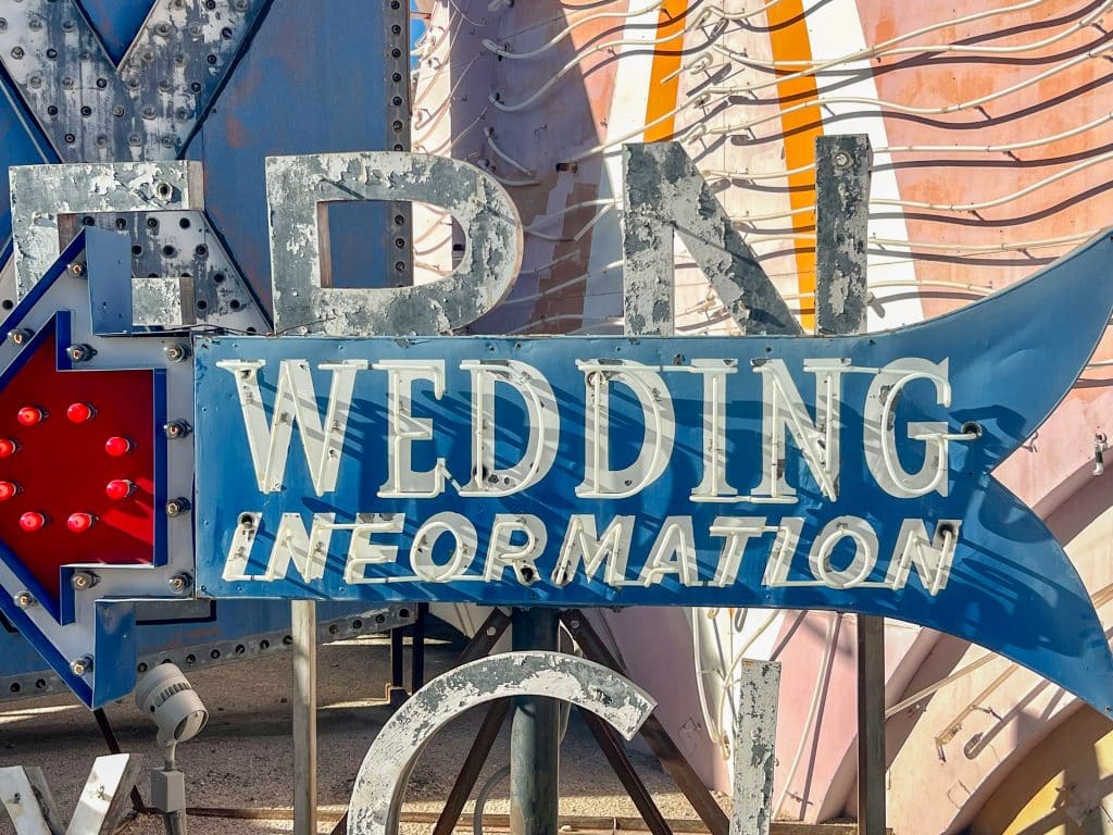 Blue metal sign in shape of arrow pointing left with words Wedding Information painted in white and containing lightbulbs. A mix of other old signage behind