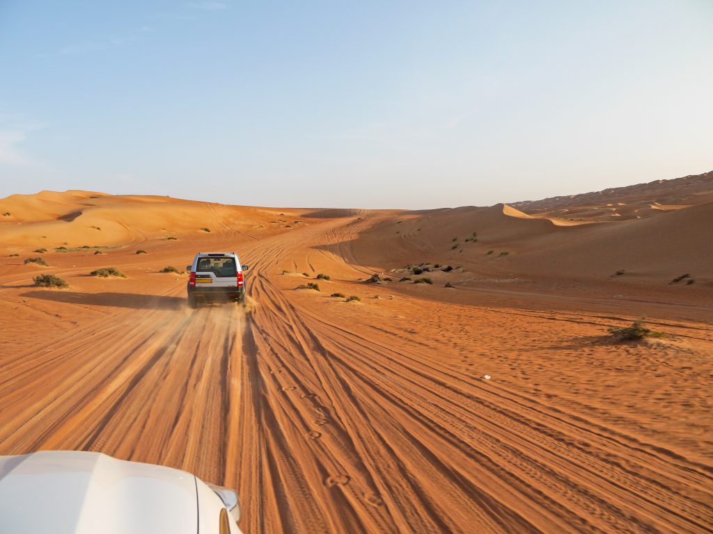 Car driving across orange sands of the desert at sundown. Can see the tyre tracks in the sand.