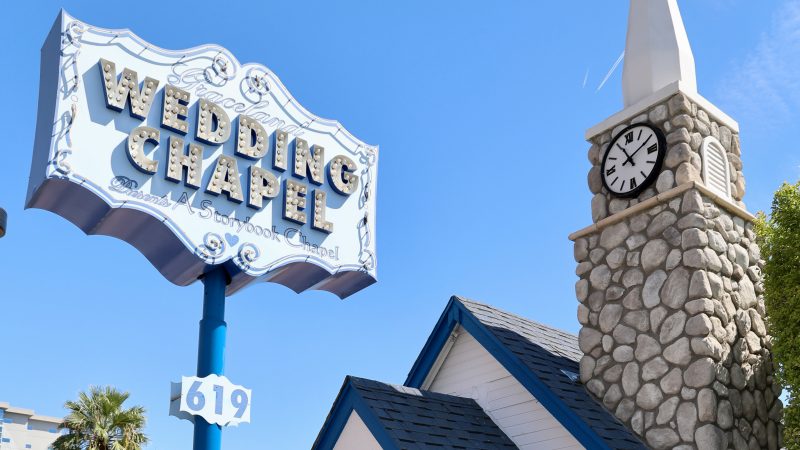 Blue sky with a small chapel on the right hand side - brick steeple with a white top and clock face, blue roofs on white wooden walls. To the left is an old fashioned sign saying Wedding Chapel on it