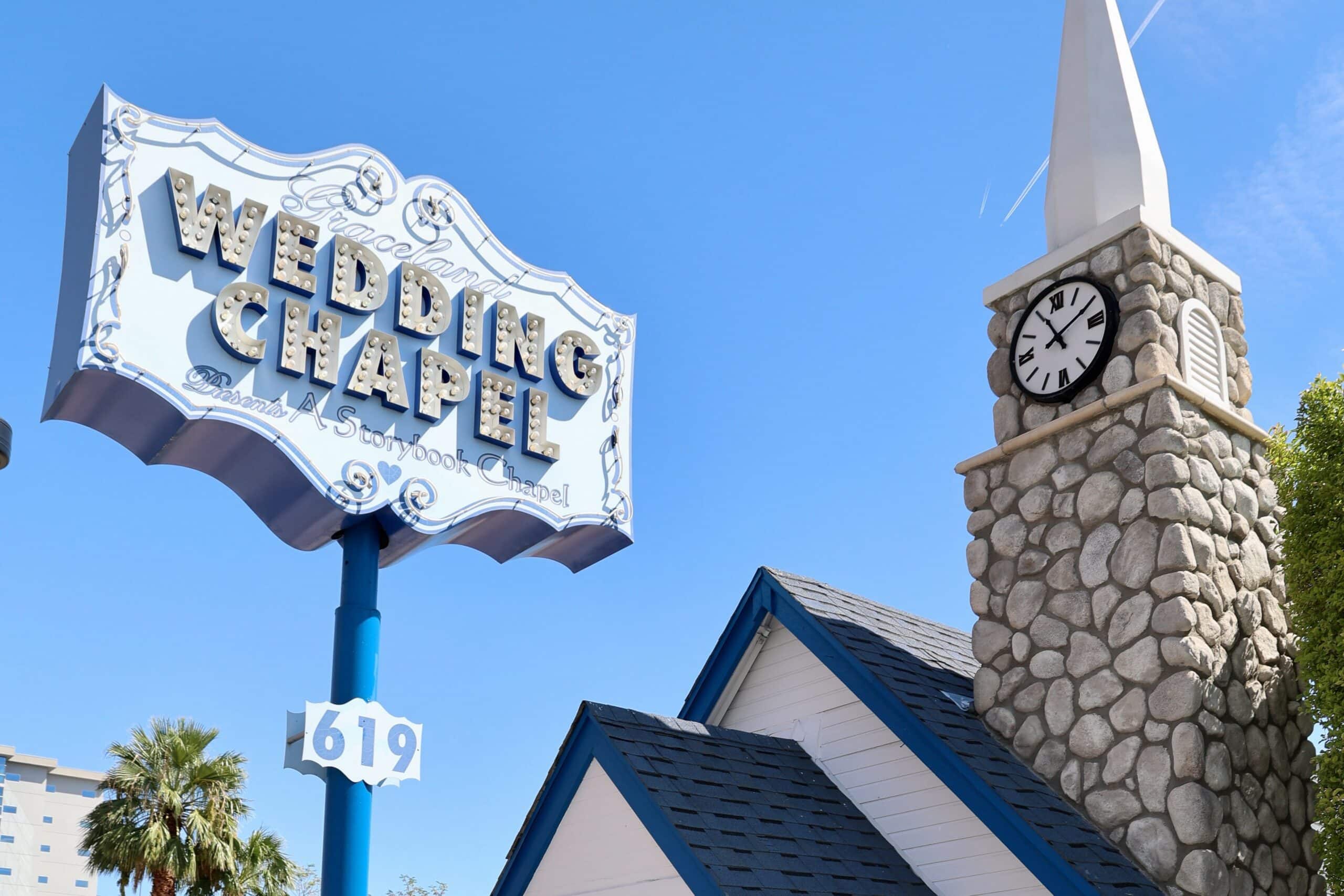 Blue sky with a small chapel on the right hand side - brick steeple with a white top and clock face, blue roofs on white wooden walls. To the left is an old fashioned sign saying Wedding Chapel on it
