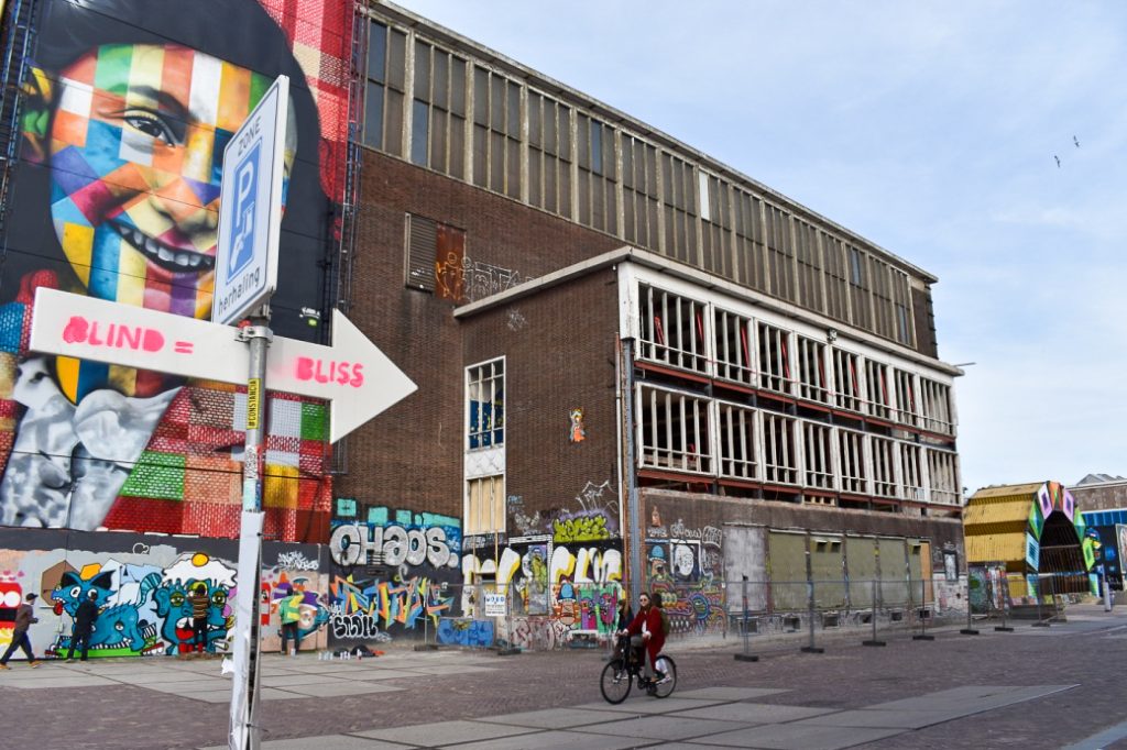 Brown factory building in centre of photo. Mural of Ann Frank painted on left hand side of the building. You can see a number of graffiti artists at the bottom, and a cyclist passing in front. White arrow sign saying Blind = Bliss on lamppost