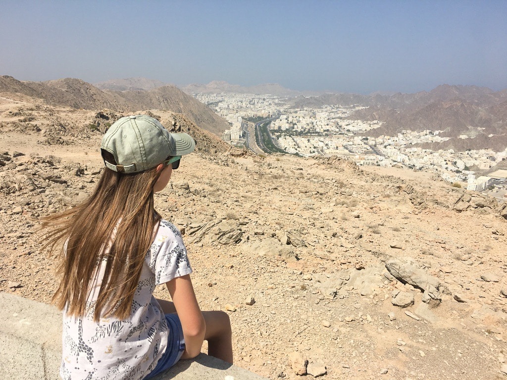Back of young girl with long brown hair wearing grey baseball hat and white t-shirt. Girl is sitting on rocks overlooking a valley with white buildings below